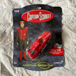 Peut inclure: Une voiture jouet rouge avec un personnage de Captain Scarlet sur le côté. La voiture a un bouton sur le dessus et indique "Try Me" et "3 Authentic Phrases (Press Button)". La voiture est emballée dans une plaquette thermoformée en plastique transparent avec un fond rouge et noir.