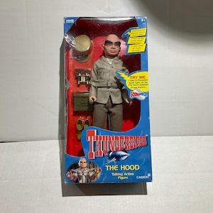 Peut inclure: Une figurine parlante "The Hood" des Thunderbirds dans sa boîte. La figurine porte un costume gris et des lunettes de soleil. Les accessoires comprennent un chapeau doré, des jumelles et une boîte verte. La boîte porte le texte "TRY ME".