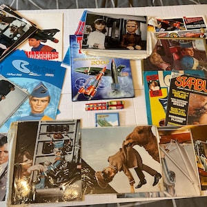 Puede incluir: Una colección de recuerdos de Thunderbirds que incluye varias revistas impresas, carteles promocionales y fotografías de personajes con marionetas de la serie de televisión. Los artículos muestran gráficos coloridos, texto e imágenes de vehículos y personajes.