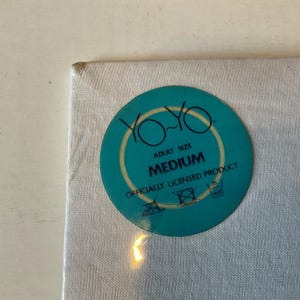 May include: A white fabric with a teal circular tag. The tag features the words "YO-YO" in a yellow font, with "ADULT SIZE" above "MEDIUM". The tag also includes washing instructions and the text "OFFICIALLY LICENSED PRODUCT".