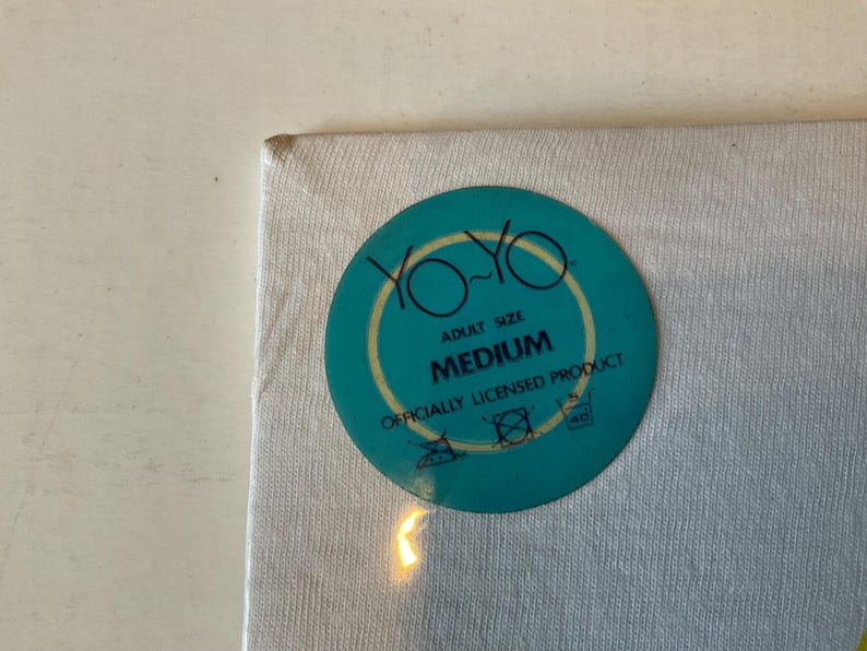 May include: A white fabric with a teal circular tag. The tag features the words "YO-YO" in a yellow font, with "ADULT SIZE" above "MEDIUM". The tag also includes washing instructions and the text "OFFICIALLY LICENSED PRODUCT".