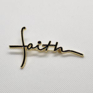Puede incluir: Pin de tono dorado con una inscripci&oacute;n de esmalte negro que dice "faith" en una fuente cursiva.