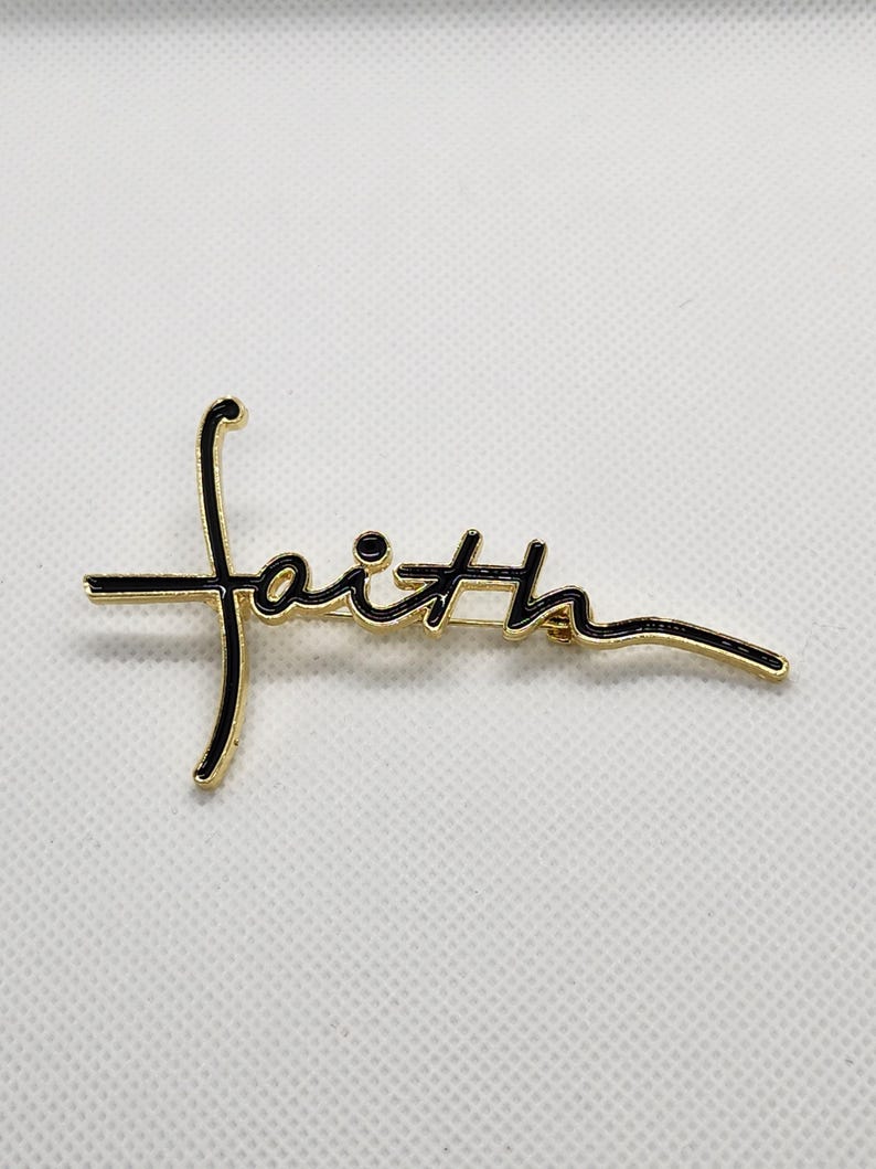 Puede incluir: Pin de tono dorado con una inscripci&oacute;n de esmalte negro que dice "faith" en una fuente cursiva.
