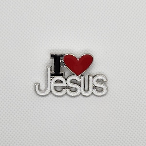 May include: A silver-colored pin with the words "I love Jesus". The letter "I" is black, the heart is red, and the word "Jesus" is in a silver font with a white outline. The pin is on a white background.