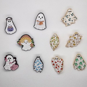 May include: Ten enamel pins featuring different ghost designs. The ghosts are white with various colored patterns and details. Some ghosts are wearing hats, holding books, or using a phone.