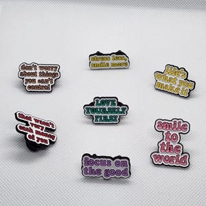 May include: Set of 7 enamel pins with motivational quotes. The pins are white with colorful text and black outlines. The quotes are: "Don't worry about things you can't control", "Stress less, smile more", "Love yourself first", "That wasn't very cash money of you", "Focus on the good", "Life's what you make it", and "Smile to the world".