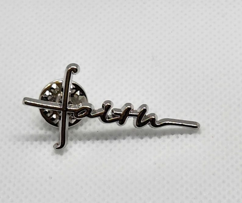 Puede incluir: Pin plateado con una cruz y la palabra "fe" escrita en cursiva.