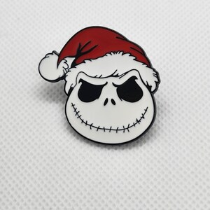May include: A pin featuring Jack Skellington from The Nightmare Before Christmas, wearing a Santa hat. The pin has a white skull face with black eyes and a stitched smile, outlined in black. The Santa hat is red with white trim.