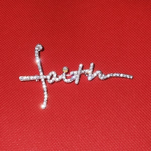 Puede incluir: Un broche plateado con la palabra "fe" escrita en diamantes de imitaci&oacute;n. El broche est&aacute; sobre un fondo rojo.