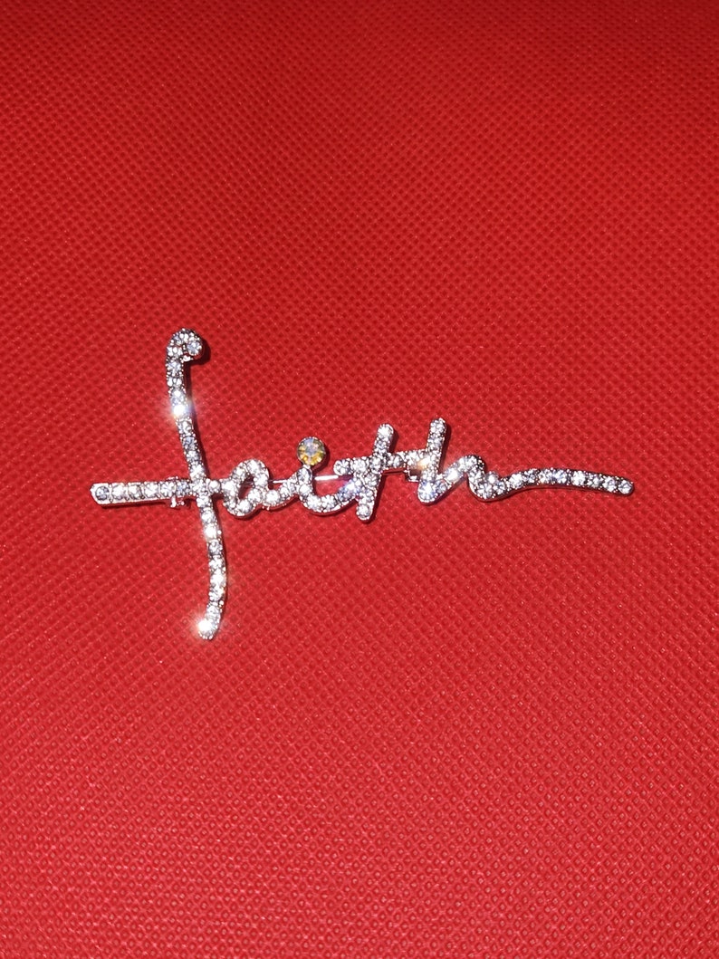 Puede incluir: Un broche plateado con la palabra "fe" escrita en diamantes de imitaci&oacute;n. El broche est&aacute; sobre un fondo rojo.