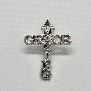 Puede incluir: Cruz de plata con la palabra "GRACE" escrita horizontalmente en la parte superior y la palabra "AMAZING" escrita verticalmente en el centro.