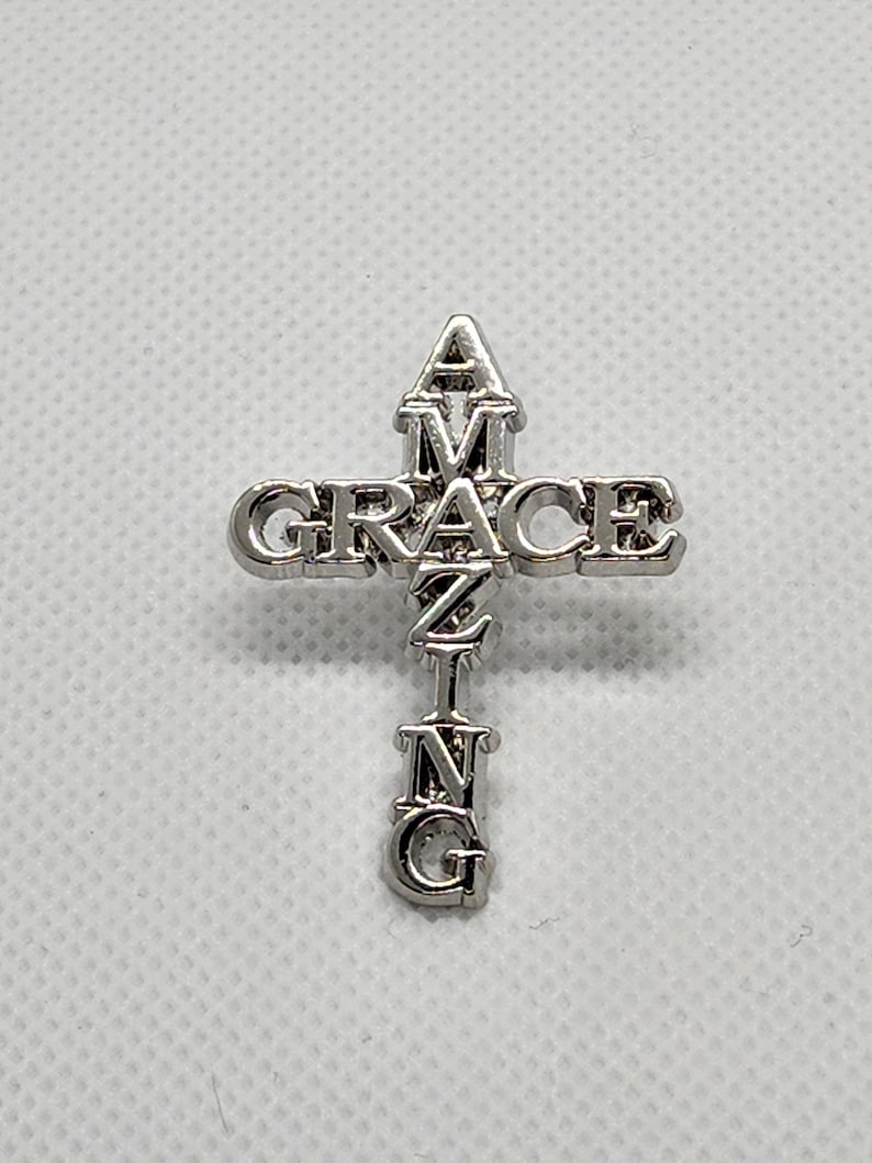Puede incluir: Cruz de plata con la palabra "GRACE" escrita horizontalmente en la parte superior y la palabra "AMAZING" escrita verticalmente en el centro.
