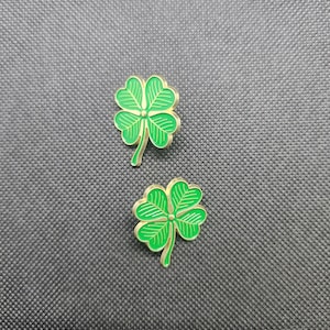May include: Two gold-colored four-leaf clover pins with green enamel. The pins are on a black textured background.