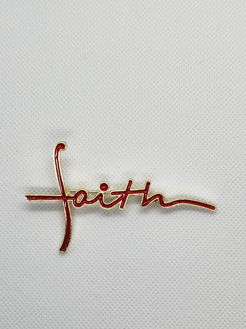 Puede incluir: Un pin de tono dorado con la palabra "fe" escrita en letras cursivas rojas. El pin presenta un dise&ntilde;o de cruz.