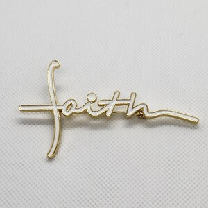 Puede incluir: Pin de tono dorado con la palabra "faith" escrita en esmalte blanco.