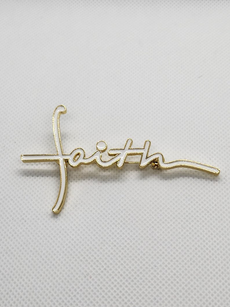 Puede incluir: Pin de tono dorado con la palabra "faith" escrita en esmalte blanco.
