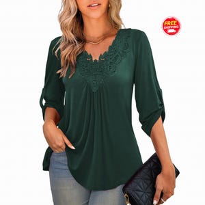 May include: A dark green blouse featuring a V-neckline with intricate lace detailing, three-quarter length sleeves with roll-tab cuffs, and a pleated front bodice. The garment is paired with light blue denim jeans and a black quilted handbag.