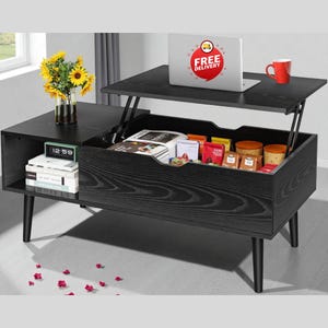 May include: A black lift-top coffee table with a storage compartment. A laptop with a "FREE DELIVERY" sticker sits on the raised top. A red mug and a vase of sunflowers are also on the table. The table has a mid-century modern design.