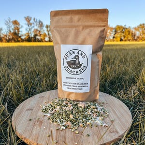 May include: A brown paper bag of "Peas and Quackers" high-protein snack mix, with a duck logo, sits on a wooden cutting board. The mix includes dried peas, shrimp, and cracked corn. The board rests on grass.