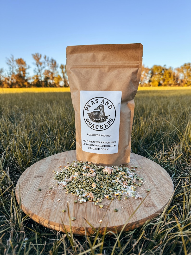 May include: A brown paper bag of "Peas and Quackers" high-protein snack mix, with a duck logo, sits on a wooden cutting board. The mix includes dried peas, shrimp, and cracked corn. The board rests on grass.