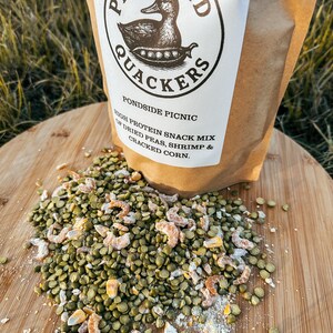 May include: A brown paper bag labeled "Peas and Quackers" with a duck illustration, containing a high-protein snack mix. The mix includes dried peas, shrimp, and cracked corn, scattered on a wooden surface.