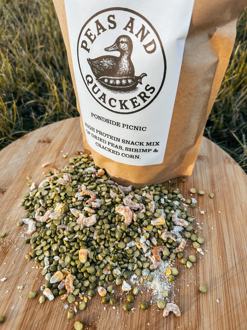 May include: A brown paper bag labeled "Peas and Quackers" with a duck illustration, containing a high-protein snack mix. The mix includes dried peas, shrimp, and cracked corn, scattered on a wooden surface.