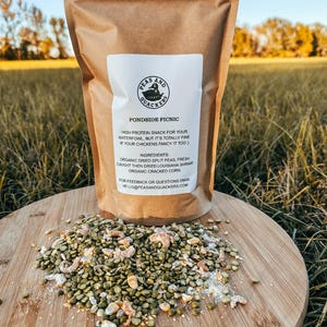 May include: A brown paper bag labeled "Peas and Quackers Pondside Picnic" with a white label. The bag sits on a wooden cutting board with a pile of green split peas, shrimp, and corn. The background is a grassy field.