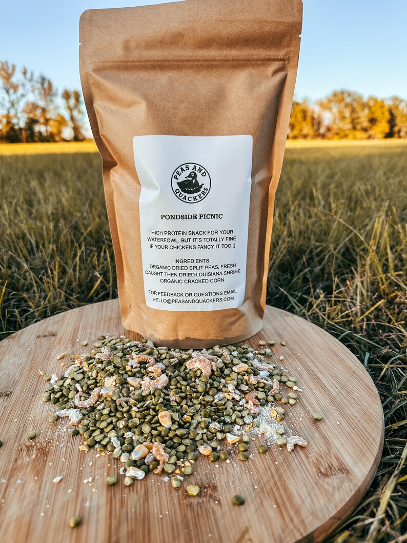 May include: A brown paper bag labeled "Peas and Quackers Pondside Picnic" with a white label. The bag sits on a wooden cutting board with a pile of green split peas, shrimp, and corn. The background is a grassy field.