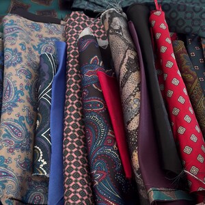 May include: A collection of rolled fabric swatches featuring various patterns including paisley, geometric shapes, and solid colors in shades of red, blue, black, brown, and tan. The assortment displays diverse textures and intricate prints.