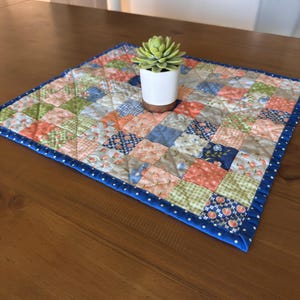 May include: A square quilted placemat featuring a patchwork design of various floral and geometric patterns in shades of peach, green, and blue, finished with a blue and white polka dot border, topped with a small succulent in a white pot.