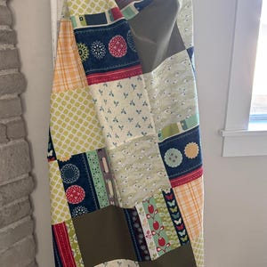 May include: A patchwork quilt featuring a variety of fabric patterns including yellow plaid, green lattice, navy blue with floral circles, olive green solid blocks, and light green fabric with small white sheep and red berry motifs.