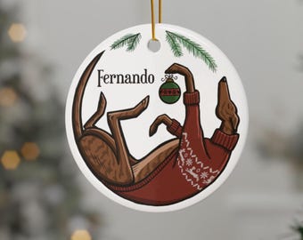 Può includere: Ornamento natalizio rotondo in ceramica. Raffigura un cane marrone con un maglione rosso con un motivo a fiocco di neve bianco, che tiene in mano una palla verde e rossa. Il nome "Fernando" è stampato sull'ornamento.