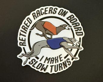 May include: White sticker with a black circle and text that reads "Retired Racers On Board I Make Slow Turns". Two greyhounds are depicted running, one wearing an orange and white racing vest and the other wearing a blue and white racing vest.