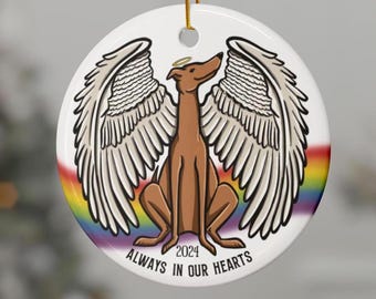 Op de afbeelding: Een witte keramische ornament met een regenboogachtergrond en een bruine hond met engelenvleugels. De hond zit met zijn hoofd omhoog gekanteld en heeft een halo boven zijn hoofd. De tekst "2024 ALWAYS IN OUR HEARTS" is onder de hond gedrukt.