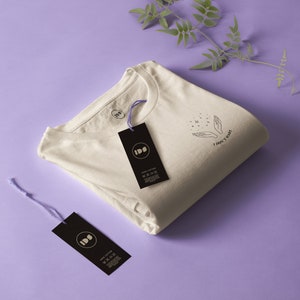 Può includere: Una maglietta bianca piegata con una stampa grafica nera che dice "I don't care" e una piccola illustrazione di mani con stelle. La maglietta ha un'etichetta nera con il testo "IDS" e "100% Cotone" stampato su di essa.