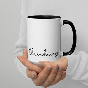 Puede incluir: Taza de café blanca con borde y asa negros. La taza tiene la palabra "thinking" escrita en cursiva negra en la parte delantera.