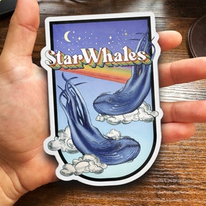 Peut inclure: Un autocollant représentant deux baleines bleues nageant dans un ciel étoilé de nuit avec un arc-en-ciel. Le texte "Star Whales" est écrit dans une police rétro.