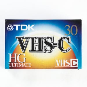 Könnte beinhalten: Ein blaues und gelbes VHS-C-Videokassettenband mit dem aufgedruckten Text "TDK", "VHS-C", "HG ULTIMATE", "VHS-C" und "30".