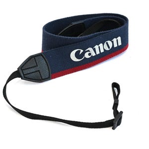 May include: A navy blue Canon camera strap with a red stripe and white lettering. The strap has a black leather connector and a black adjustable strap with a plastic buckle. The strap is designed to be worn around the neck.