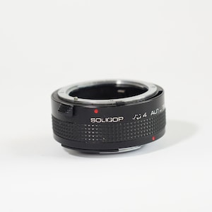 May include: A black and silver camera lens adapter with the text "Soligor 1/34 Auto" printed on the side.