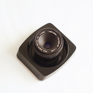 Könnte beinhalten: Ein schwarzes Kameraobjektiv mit dem Text "FUJI PHOTO OPTICAL CO.  FUJINON-E 1:1.9/50  LENS-JAPAN" um das Objektiv herum.