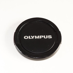 Puede incluir: Una tapa de lente de cámara negra con la palabra "OLYMPUS" impresa en letras plateadas.