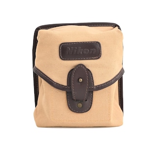 May include: A rectangular camera bag made of tan canvas with dark brown leather trim. The front flap features a dark brown leather patch embossed with the word Nikon and a central leather strap closure with a metal snap button fastener.