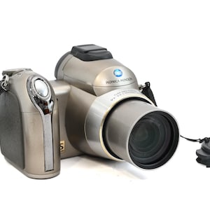 May include: A silver Konica Minolta digital camera with a black strap and lens cap. The camera has a large lens and a built-in flash. The Konica Minolta logo is visible on the top of the camera.
