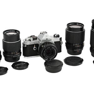 Könnte beinhalten: Eine silberne und schwarze Pentax-Kamera mit mehreren schwarzen Objektiven und Objektivdeckeln. Die Objektive sind unterschiedlich groß und um die Kamera herum angeordnet. Die Ausrüstung befindet sich auf weißem Hintergrund. Fotoausrüstung.