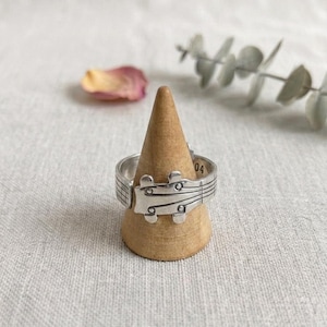 Könnte beinhalten: Ein silberfarbener Metallring mit einem Design, das an die Kopfplatte eines Saiteninstruments erinnert, komplett mit Stimmwirbeldetails und eingravierten Notenlinien auf dem Ringband. Im Inneren des Ringbandes ist die Zahl 04 zu sehen.