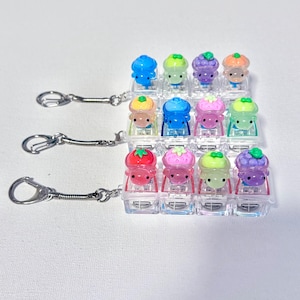May include: A collection of colorful miniature ice cream cone keychains. Each keychain features a different colored ice cream cone with a smiling face, including blue, green, purple, and orange. The keychains are attached to silver-colored metal rings.
