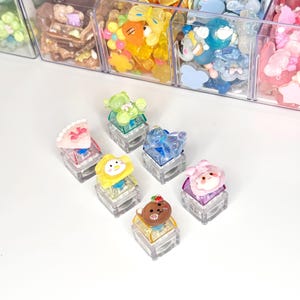 Könnte beinhalten: Eine Sammlung von sechs dekorativen mechanischen Tastaturkappen mit verschiedenen bunten Harzfiguren, darunter ein grüner Bär, ein blaues Tier, ein gelbes Küken, eine rosa Figur und ein brauner Bär, angeordnet vor einem durchsichtigen Kunststoff-Organizer.