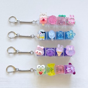 May include: Four rows of colourful keychains with various charms. Each keychain has a silver clasp and a spring coil. Charms include ice cream, a bunny, a bear, and a frog. The charms are in a variety of colours, including pink, blue, and purple.