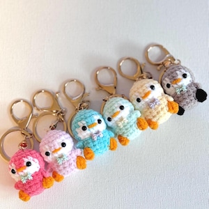 Puede incluir: Una colección de seis llaveros de pingüinos tejidos a ganchillo en varios colores, incluyendo rosa, morado claro, azul, verde menta, amarillo y gris. Cada pequeño pingüino de peluche tiene ojos de cuentas negras, pico naranja, pies naranjas y un pequeño lazo.